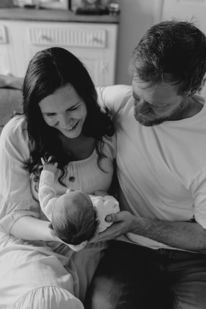 Black and white image of new parents holding their newborn baby and looking at him. Parents are close to each other and showing affection. Newborn photo shoot at home. Intimate newborn photo session. Ewa Jones Photography