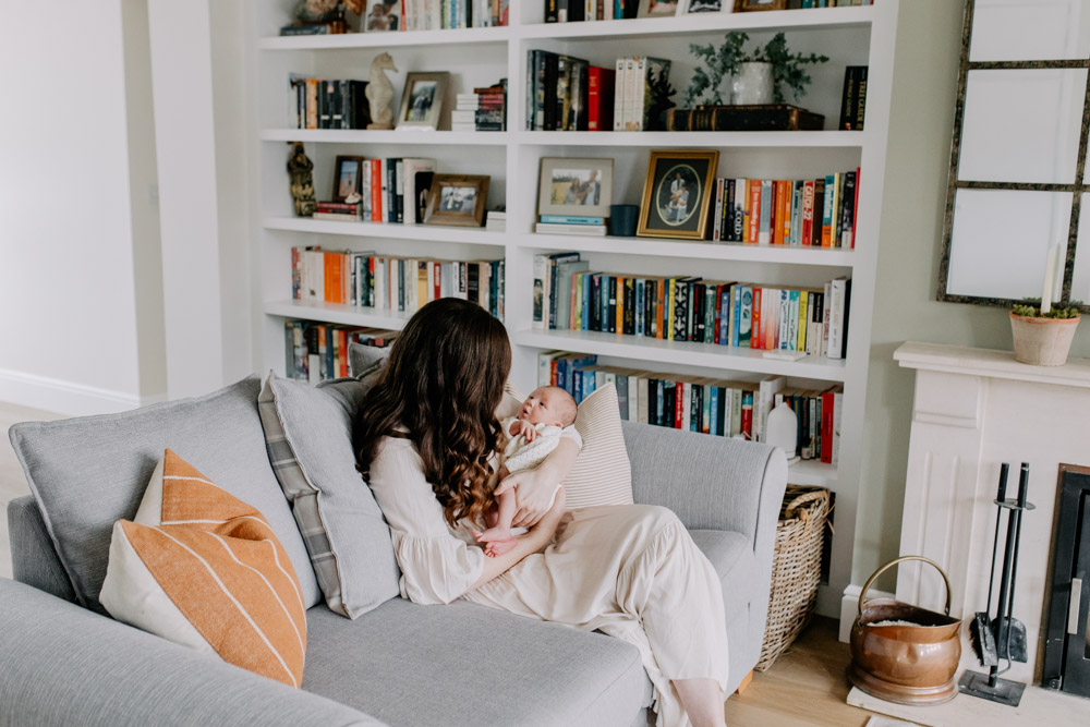 Mum is sitting holding her newborn baby and looking at baby. lovely house interior for the newborn photo session. Ewa jones Photography