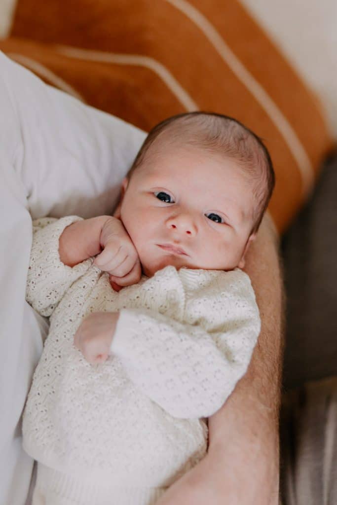 Little baby boy is being held by his dad and looking at the camera. In home newborn photo shoot at home. Newborn photo session in Basingstoke. Hampshire newborn photographer. Ewa Jones Photography