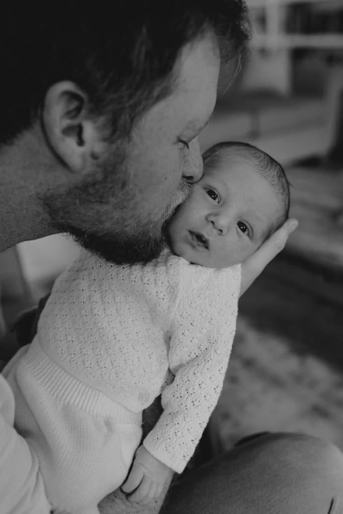 Dad is kissing his baby boy on the cheek. Newborn baby is looking at the camera. In home newborn photo session. Newborn photographer in Hampshire. Hampshire photographer. Ewa Jones Photography
