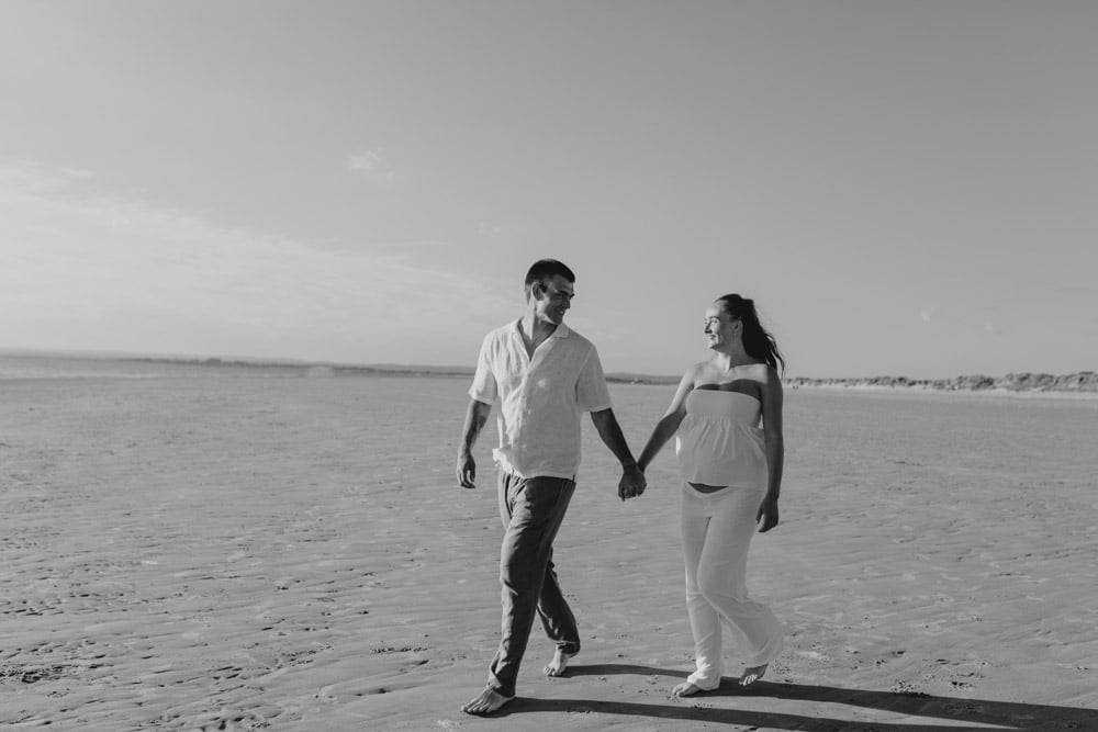 Parents to be are walking on the beach and looking and smiling at each other. lovely black and white natural image of maternity photo session. Maternity photographer in Hampshire. Ewa Jones Photography