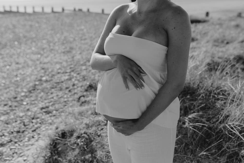 black and white image of mum holding her bump. Mum is wearing white top and trousers. lovely spring evening. Maternity photo shoot in Hampshire. Hampshire photographer. Ewa Jones Photography
