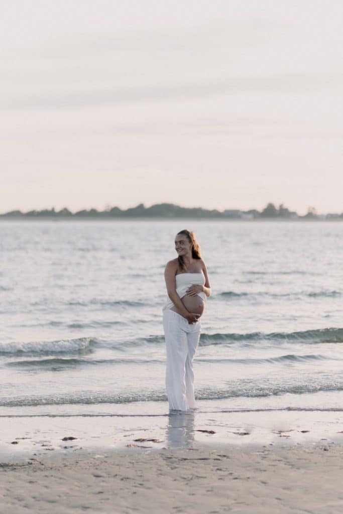 Mum to be is standing in the water and looking away. Maternity shoot at the beach. Gentle touches at the bump. Maternity shoot. Ewa Jones Photography