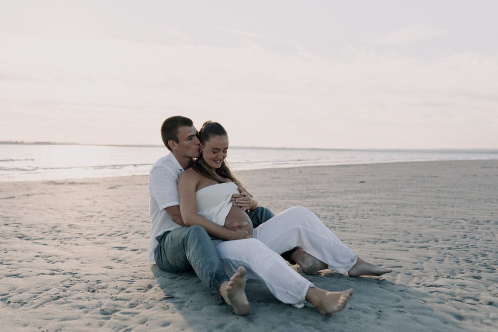 Parents to be are sitting at the beach and dad to be is kissing her partner. Spring photo shoot at the beach. maternity photo shoot. Hampshire photographer. Ewa Jones Photography