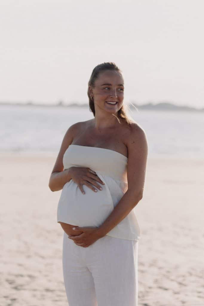 Mum to be is holding her baby bump and looking away. She is wearing white top and white trousers. maternity photo shoot. Hampshire maternity photographer. Ewa Jones Photography