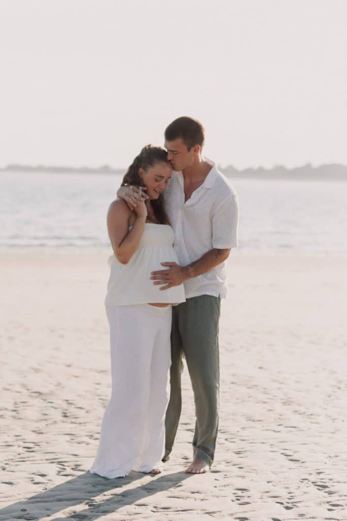Parents to be are standing at the beach. Dad is kissing the mum on her forehead. Lovely maternity photo shoot. Hampshire photographer. Ewa Jones Photography