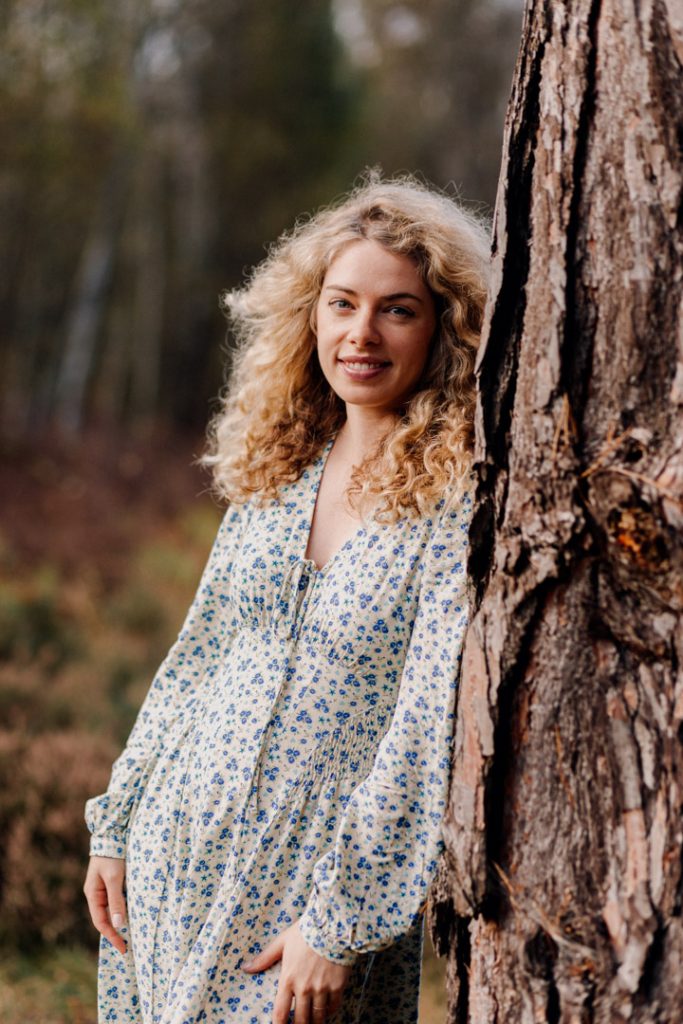 Female in white dress with blue flowers is looking at the photographer. she is standing and leaning against the tree. female photoshoot in nature. Basingstoke photographer. Ewa Jones Photography