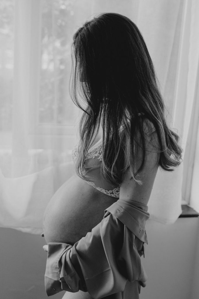 Intimate photo shoot. what to wear for maternity photo session. Mum to be is standing next to the window and looking down on baby bump. Maternity photo shoot in London. Hampshire maternity photographer. Ewa Jones Photography
