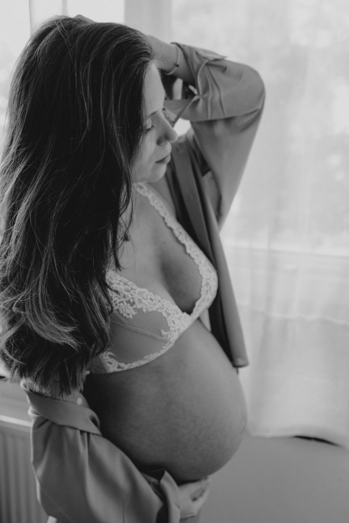 Baby bump photoshoot. Hampshire photographer. Black and white image of mum touching her hair and bump. Intimate in home photo shoot. Maternity photographer.