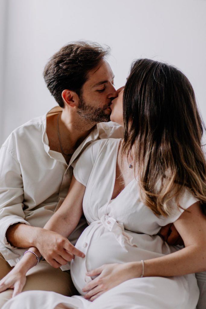 Expecting parents are kissing and sitting on the sofa. Intimate and natural maternity photo session at home. London maternity photo shoot. Hampshire photographer. Ewa Jones Photography
