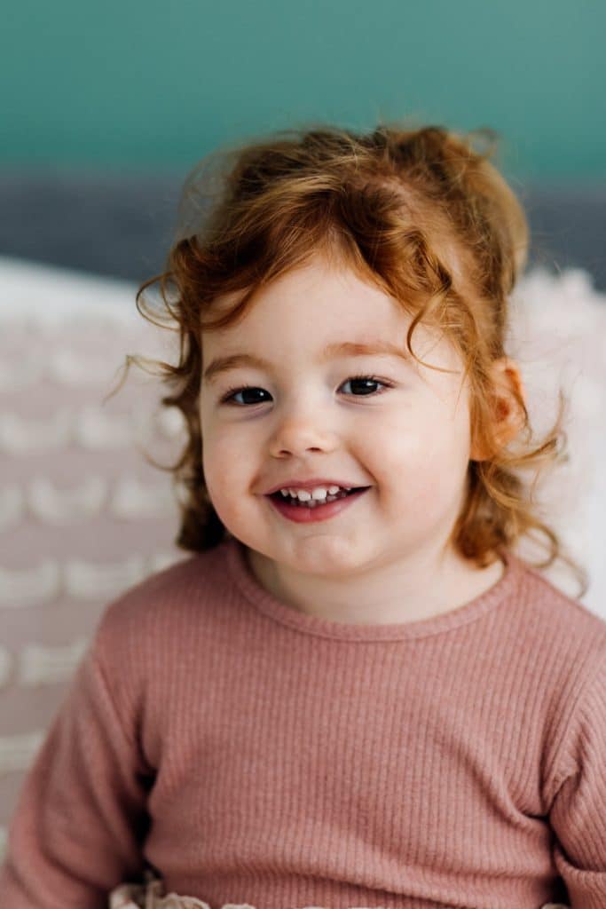 Little girl is sitting on the bed and looking at the photographer. She has lovely ginger hair. Newborn photoshoot in hampshire. Newborn photo session in Hampshire. Ewa Jones Photography