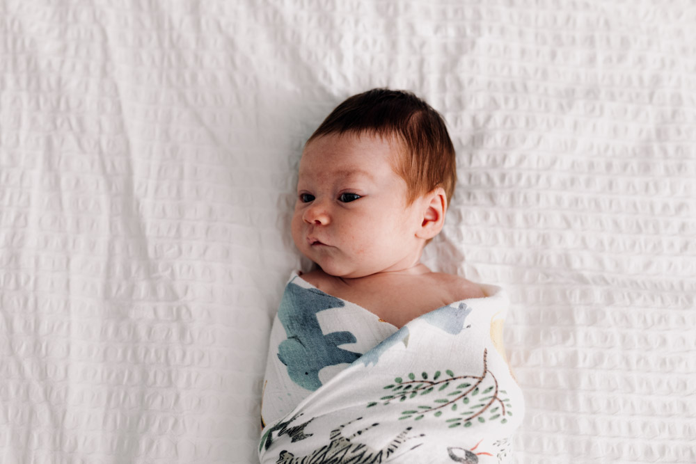 Newborn baby girl is wrapped in the muslin and laying on the bed. She is looking to the side. Natural newborn session. Newborn photographer in Hampshire. Ewa Jones Photography