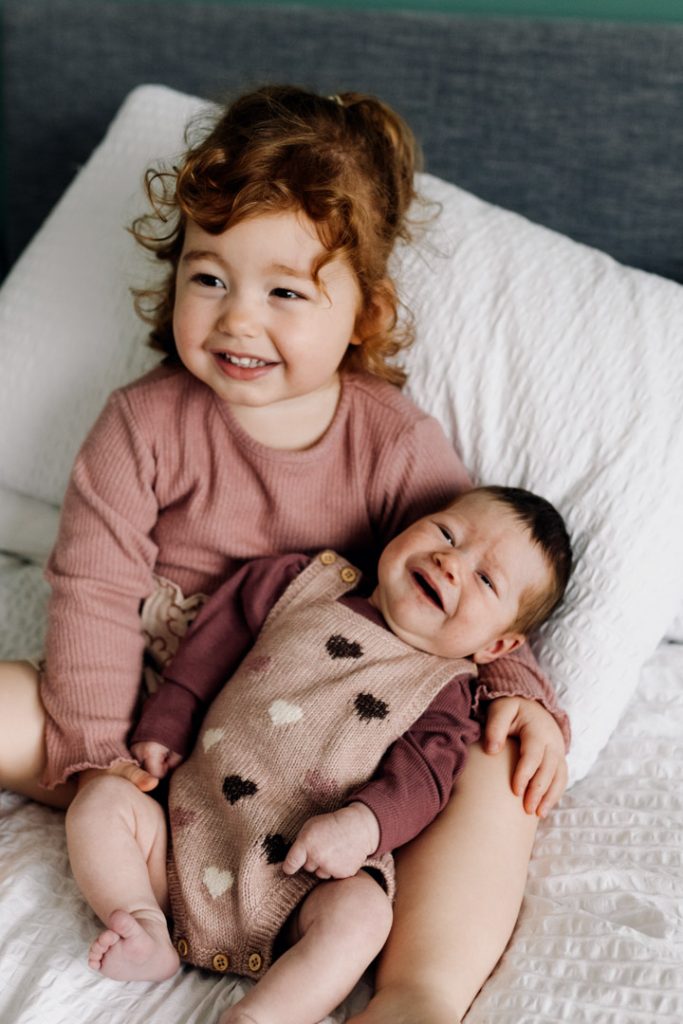 Toddler girl is holding her baby sister in her arms. Baby sister is smiling. In-home natural newborn photo session. Hampshire photographer. Ewa Jones Photography