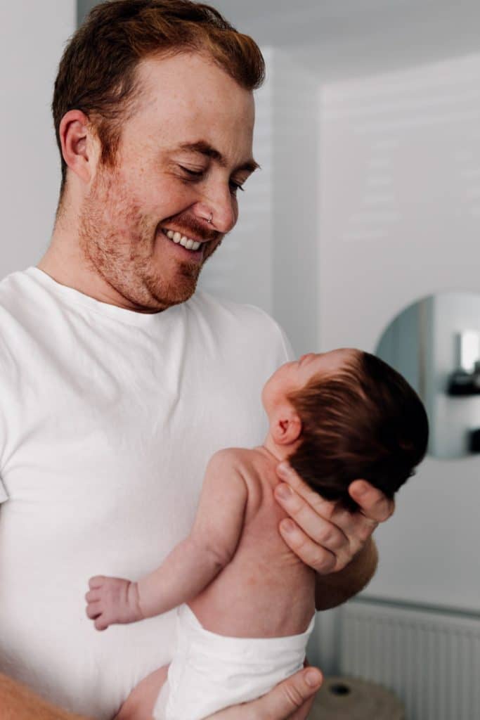 Dad is holding newborn baby in his arms and smiling to the baby. In-home natural newborn sessions. Newborn photo shoot. Newborn photographer in Basingstoke. Ewa Jones Photography