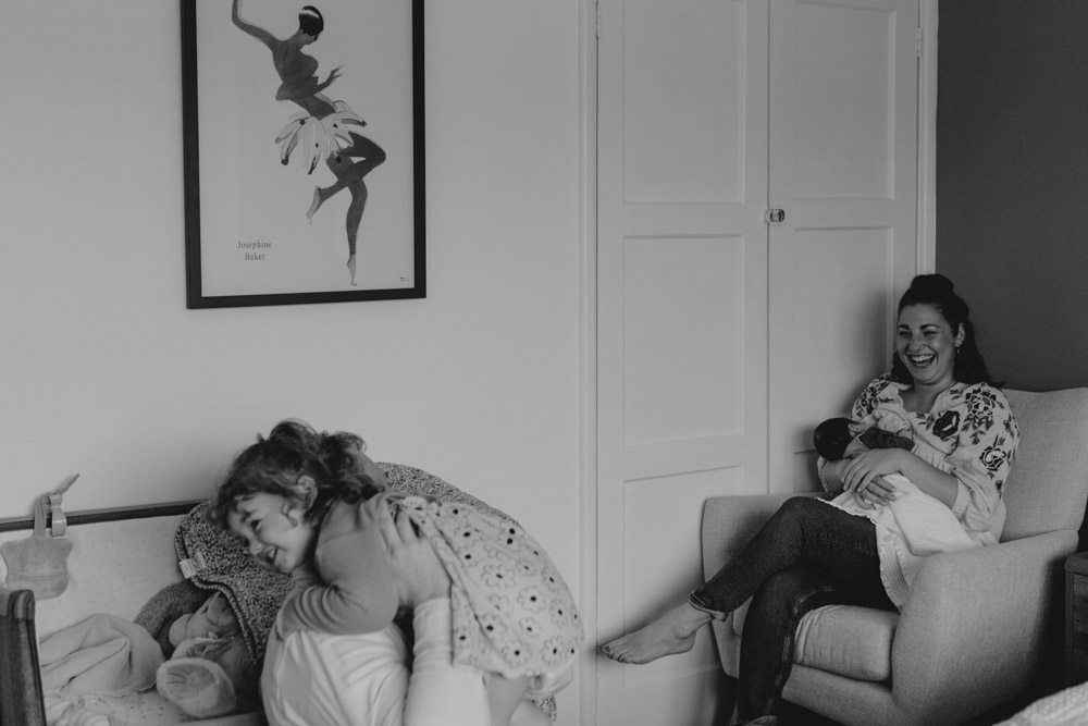Mum is sitting on the chair feeding newborn baby girl and dad is cuddling their toddler. Natural moment during in-home newborn photo session. Hampshire photographer. Ewa Jones Photography