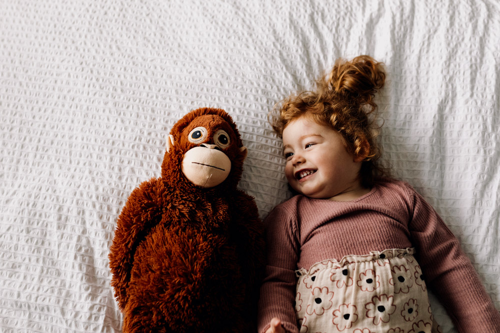 Toddler girl is laying on the bed next to a big monkey. Fun photoshoot at home. Hampshire photographer. Ewa Jones Photography