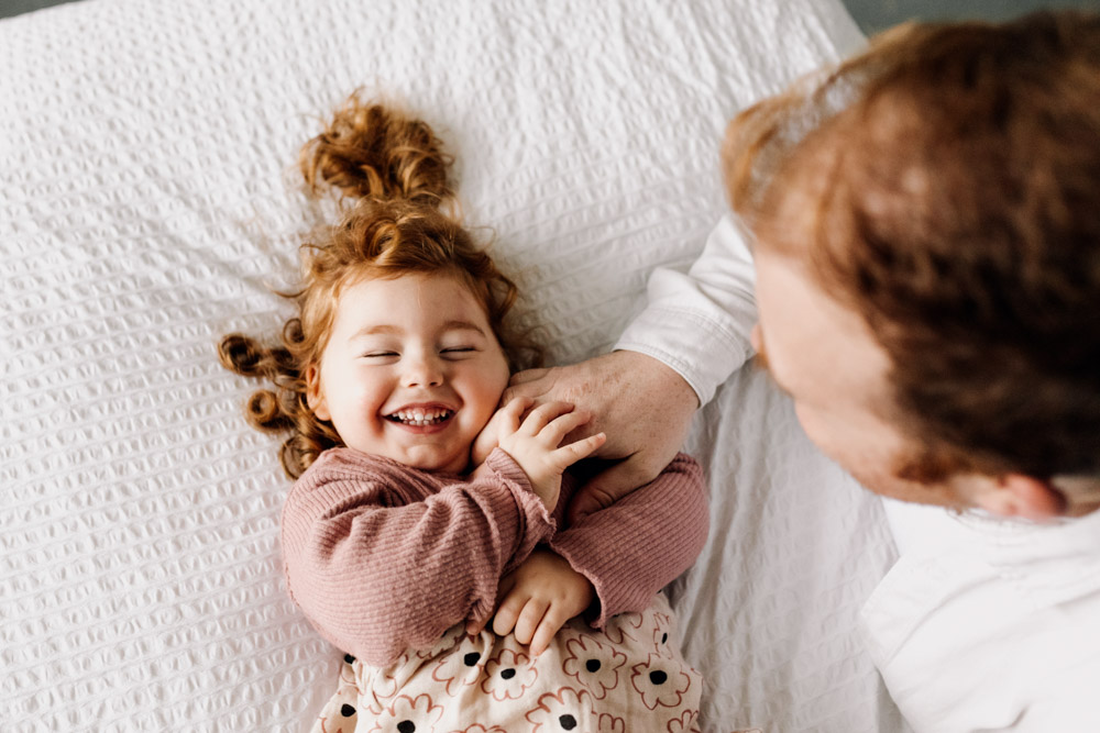 Toddler girl is being ticked on the bed y dad. Natural in home family photo session. Hampshire photographer. Ewa Jones Photography