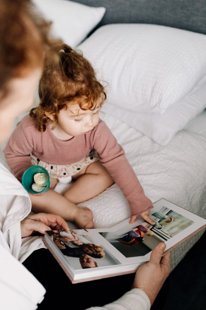 Girl is sitting on the bed and looking through an album with her dad. newborn photo session in home filled with fun and laughter. Ewa Jones Photography