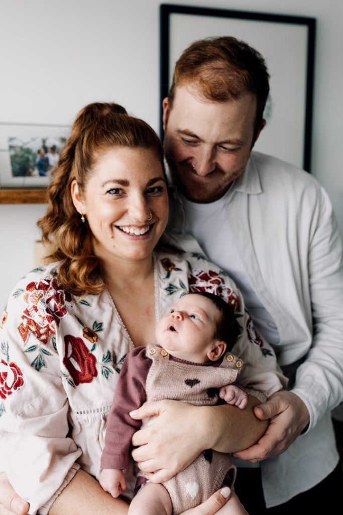 mum is holding newborn baby in her arms and dad is cuddling from behind. dad is lovingly looking at his partner. In home natural newborn photo session. Ewa Jones Photography