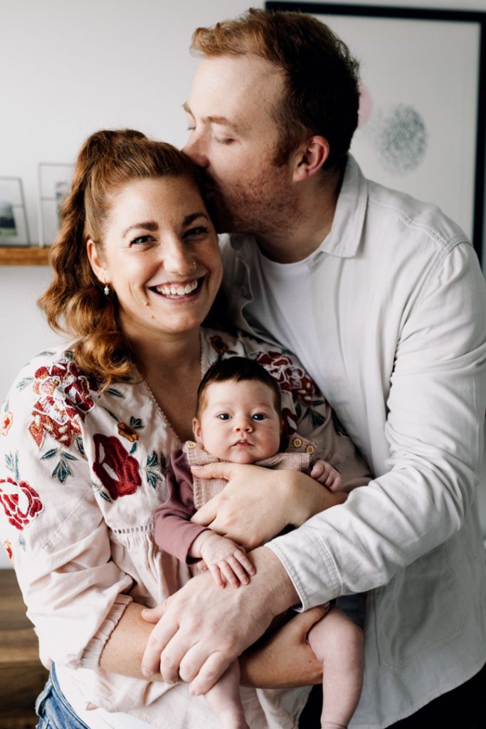 Mum is holding newborn girl and dad is cuddling and kissing mum. natural newborn photo session pose. Hampshire newborn photographer. Ewa Jones Photography
