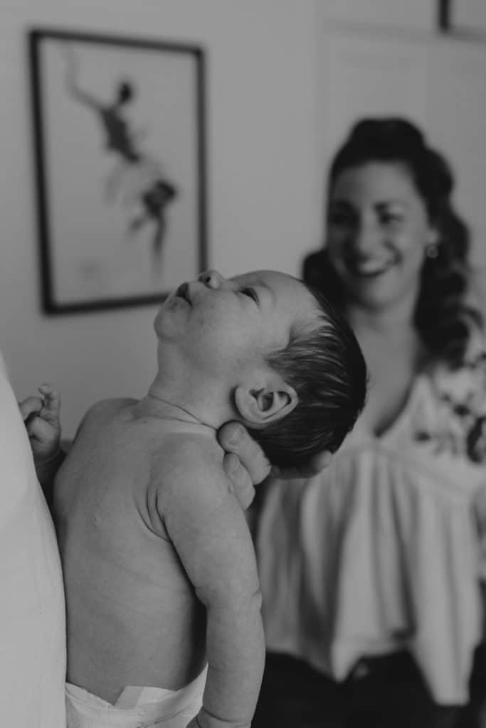 Newborn baby is being held by a dad and mum is smiling in the background. Newborn photo session in home. Natural newborn photography. Newborn photo shoot in Hampshire. Ewa Jones Photography