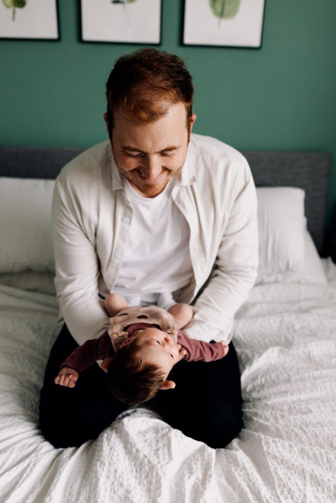 Dad is kneeling on the bed and holding his newborn baby girl in his arms. Newborn photo shoot at home. Ewa Jones Photography