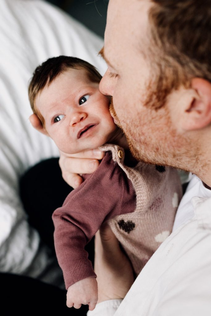Dad is kissing newborn baby on her cheek. natural newborn poses at home with dad. Hampshire newborn photo shoot. Ewa Jones Photography