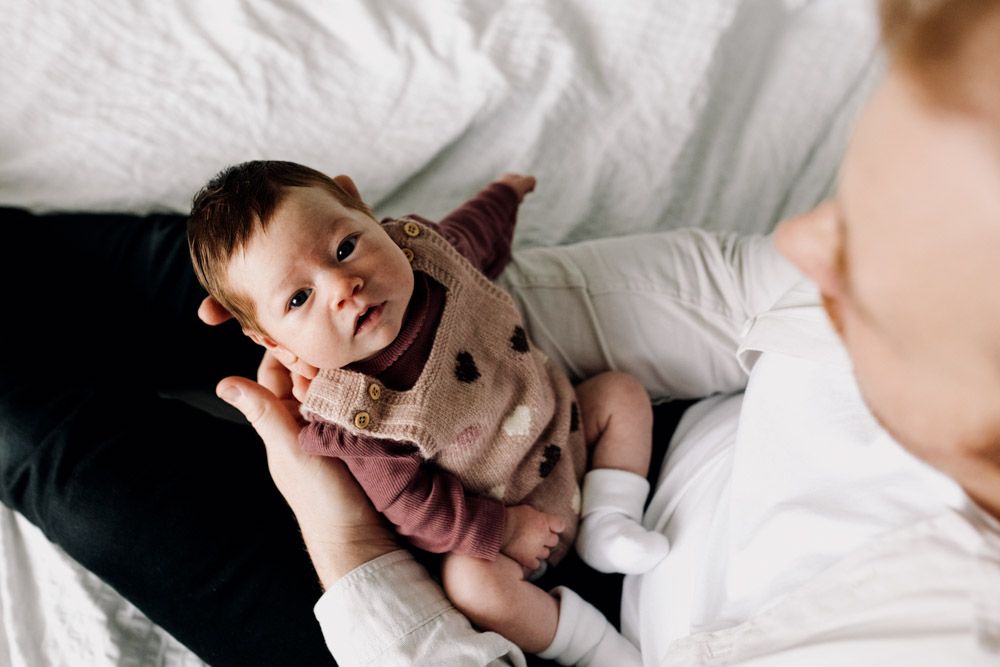 Dad is holding newborn baby girl and newborn baby is looking up at the camera. Newborn photo shoot at home. Hampshire photographer. Ewa Jones Photography
