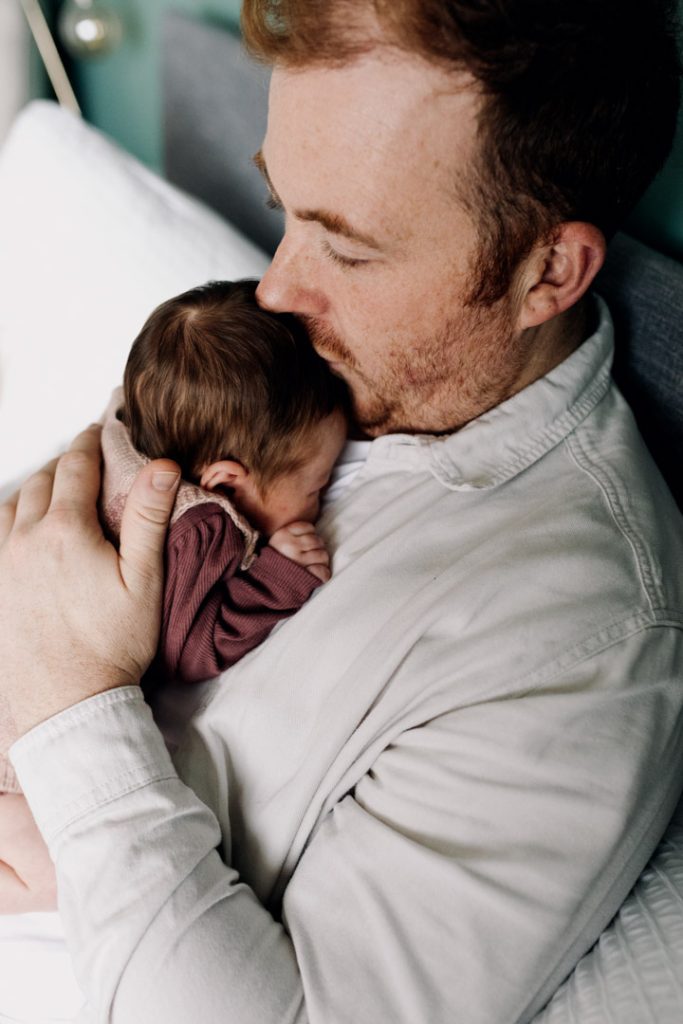 Dad is kissing newborn baby and holding her in his arms. natural unposed newborn session at home. Ewa Jones Photography