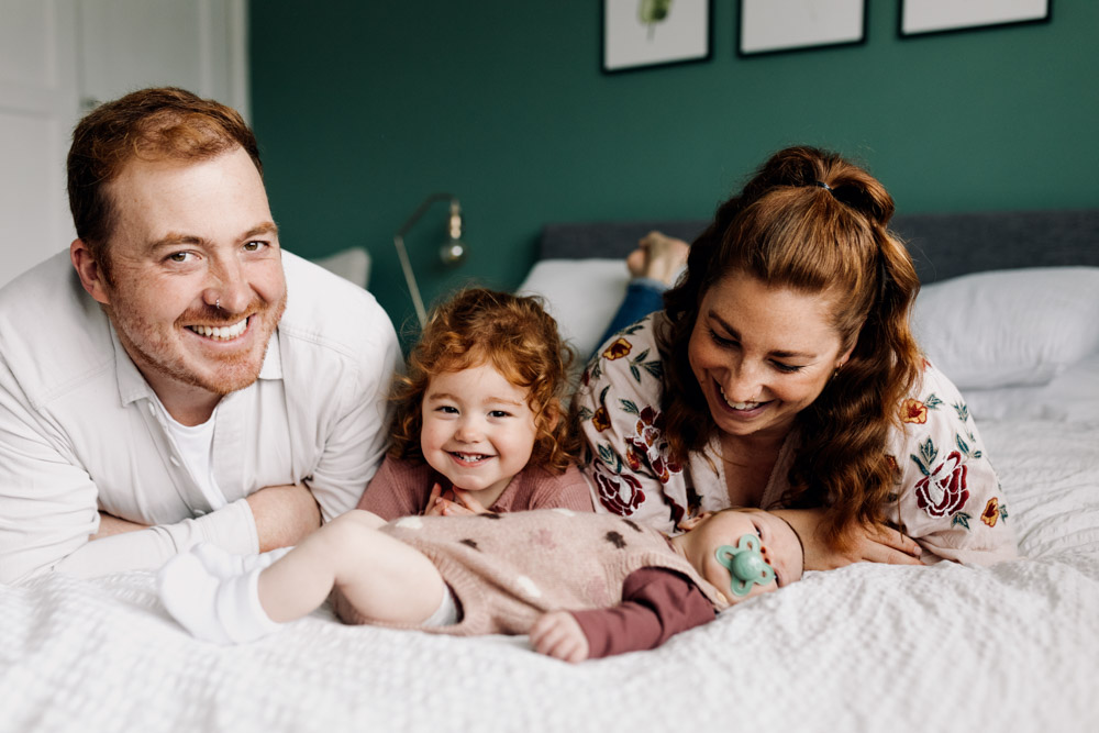 family is laying on the bed and smiling. newborn photo session at home with a toddler and newborn baby. Ewa Jones Photography