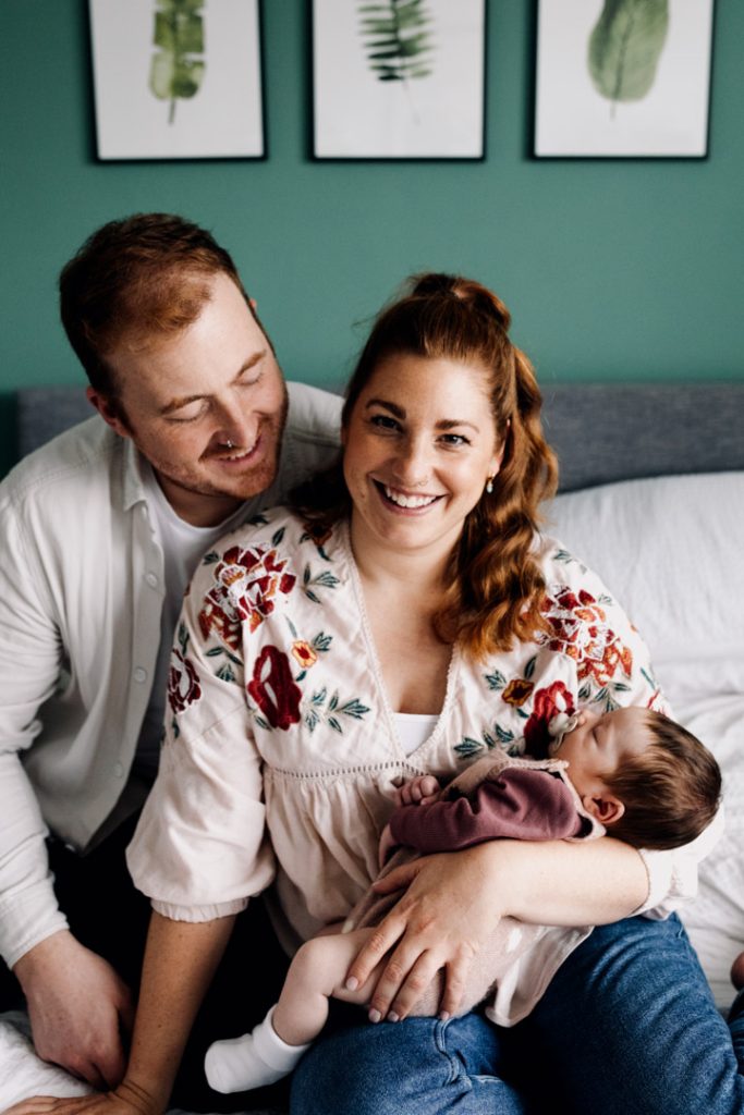 Mum is sitting on the bed and smiling. dad is looking at the baby. Hampshire newborn photoshoot. Hampshire photographer. Ewa Jones Photography