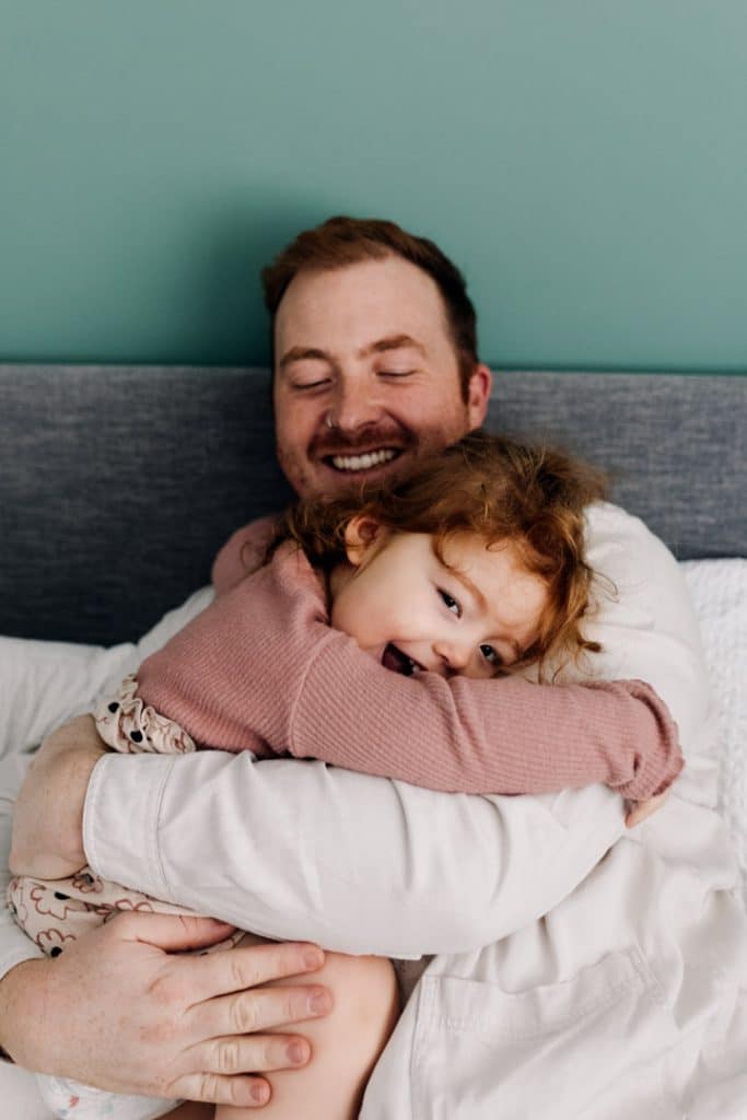 Dad is sitting on the bed and cuddling his toddler girl who is smiling. natural in-home family photo session. Newborn photographer in Hampshire. Ewa Jones Photography