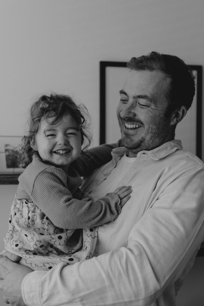 Black and white image of dad holding his little girl and smiling to her. Girl is laughing too. Family photographer in Hampshire. Newborn photo shoot. Ewa Jones Photography