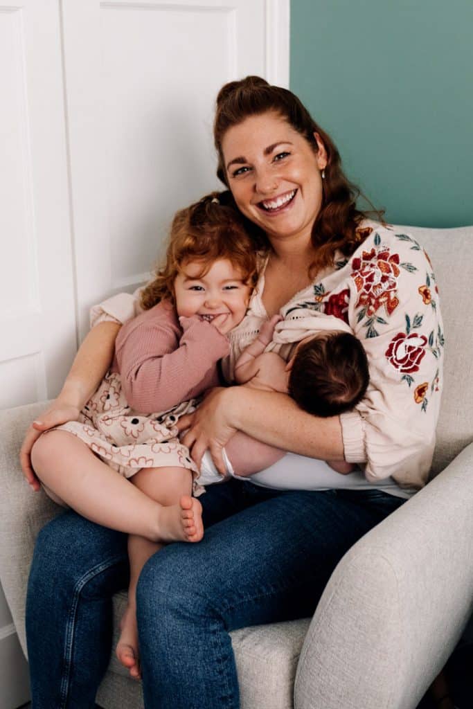 Mum is sitting on the chair and holding her newborn baby and her toddler girl in her arms. In-home newborn session with a toddler. Natural newborn photo shoot. Ewa Jones Photography