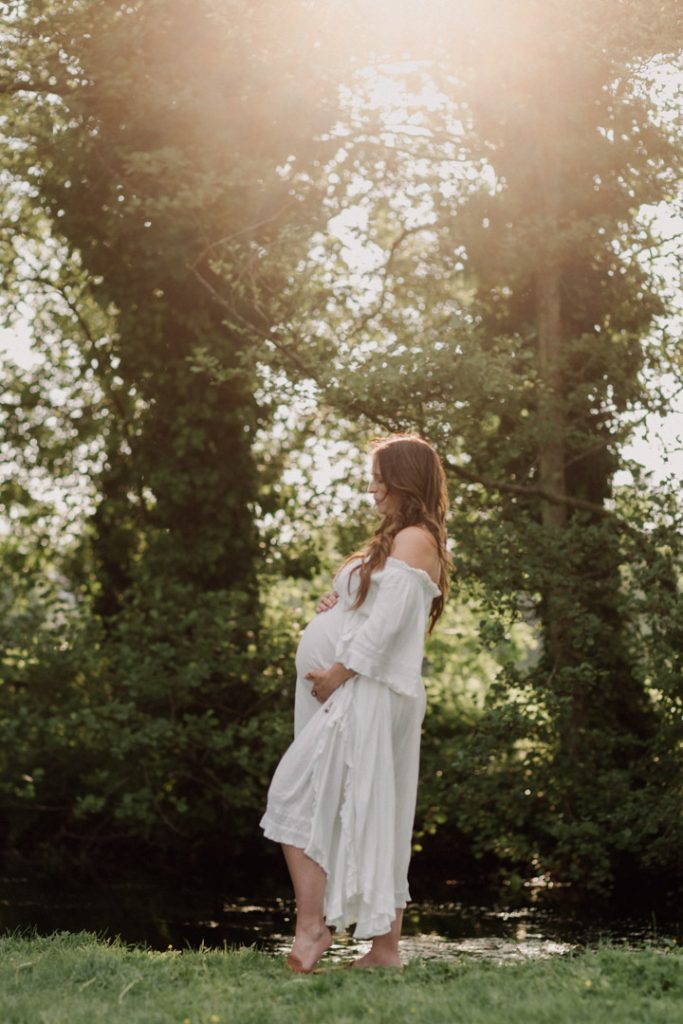 mum is standing on the grass and looking at her bump. She is wearing beautiful flowy dress showing off her bump. Golden sun is peaking through the trees. Outdoor maternity photo session in Winchester. Hampshire photographer. Ewa Jones Photographer