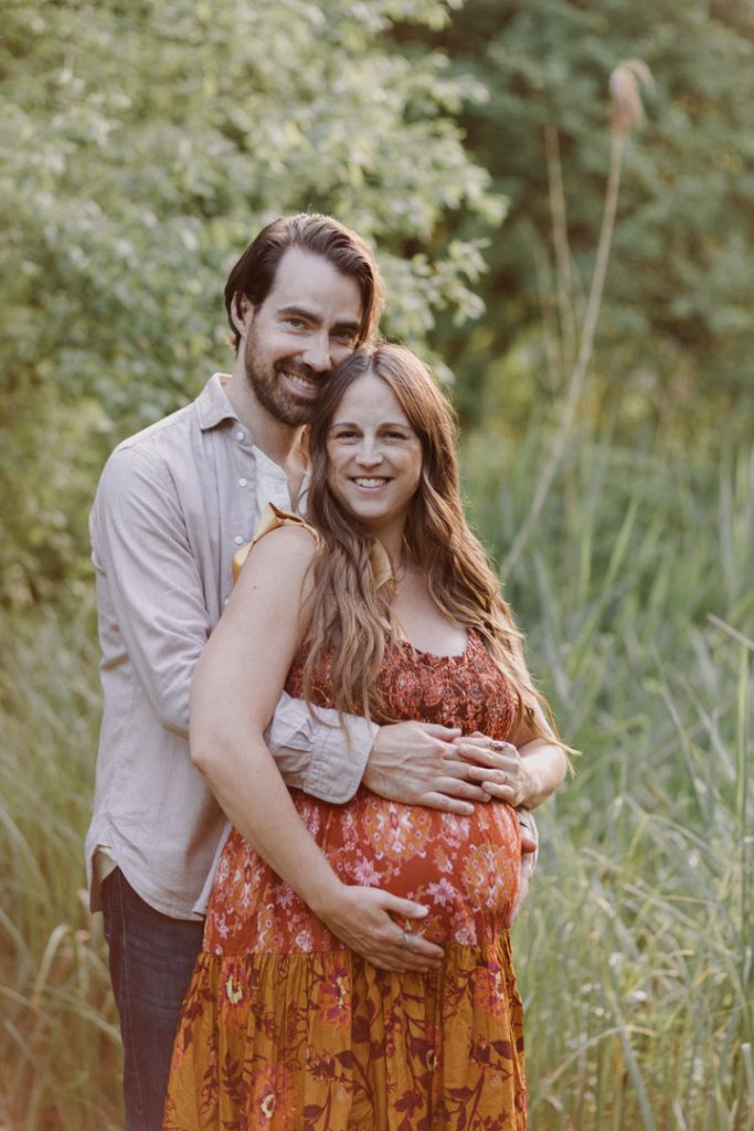 mum and dad to be are cuddling and looking at the camera. they are standing in front of lovely green foliage which is lit by golden hour. Basingstoke photographer. Ewa Jones Photographer