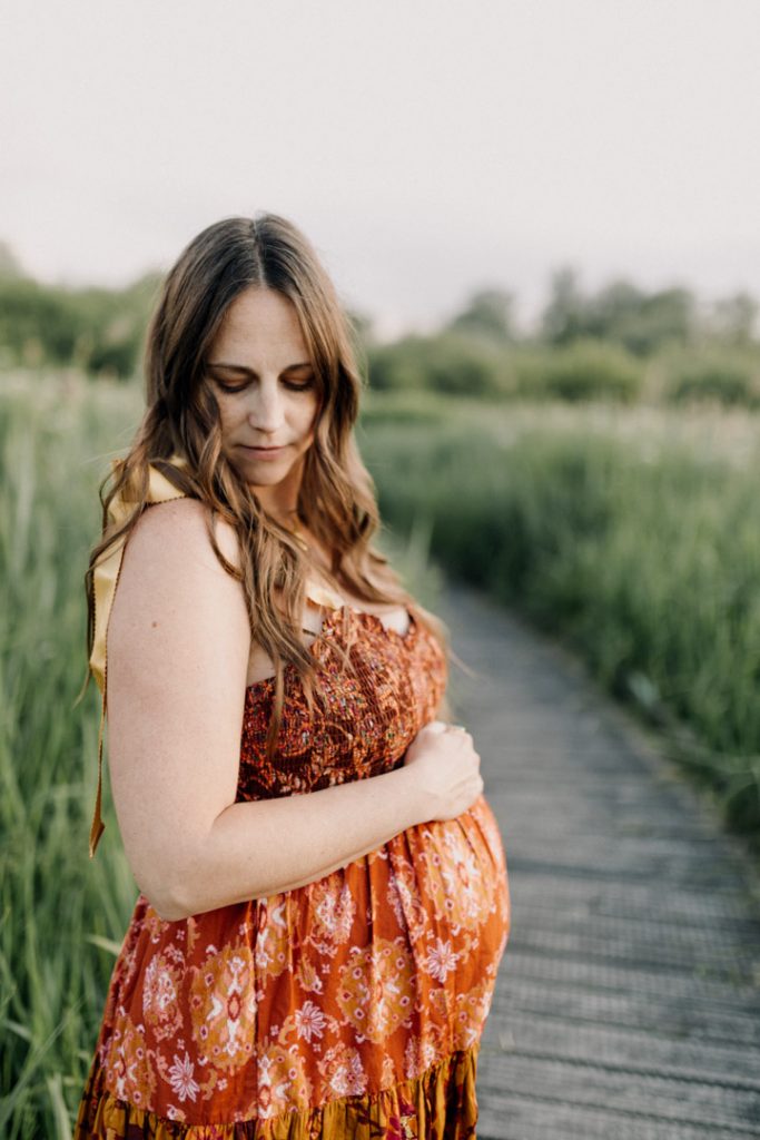Mum is standing and looking over her shoulder. she is wearing beautiful flowy dress. Maternity outdoor photo session in Hampshire. Ewa Jones Photographer