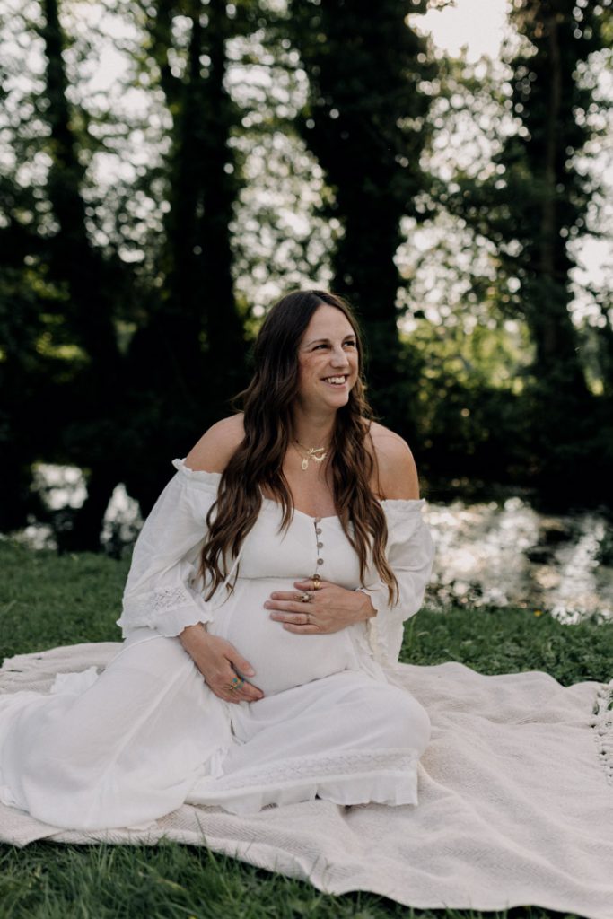mum is sitting on the blanket and holding baby bump. there are nice trees and sun in the background. Maternity photo session during golden hour. How to pose during maternity shoot. Ewa Jones Photographer