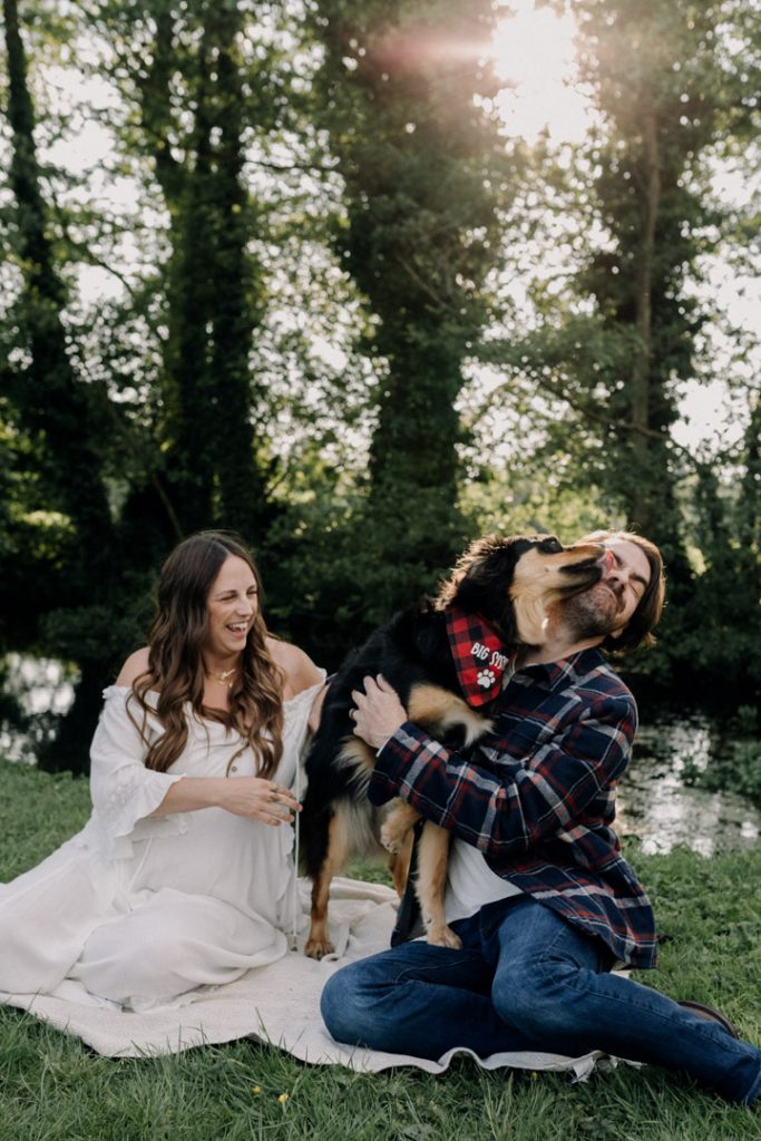 Lovely natural photo of mum to be and dad sitting on the blanket. Their dog is licking the owner. and mum is laughing. Natural bump photo shoot. Hampshire based family photographer. Ewa Jones Photographer
