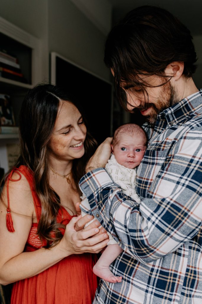 Baby is being held by dad and baby is looking out. mum and dad are happy and looking down at their baby. Newborn photo session in Hampshire. Basingstoke family photographer. Ewa Jones Photography