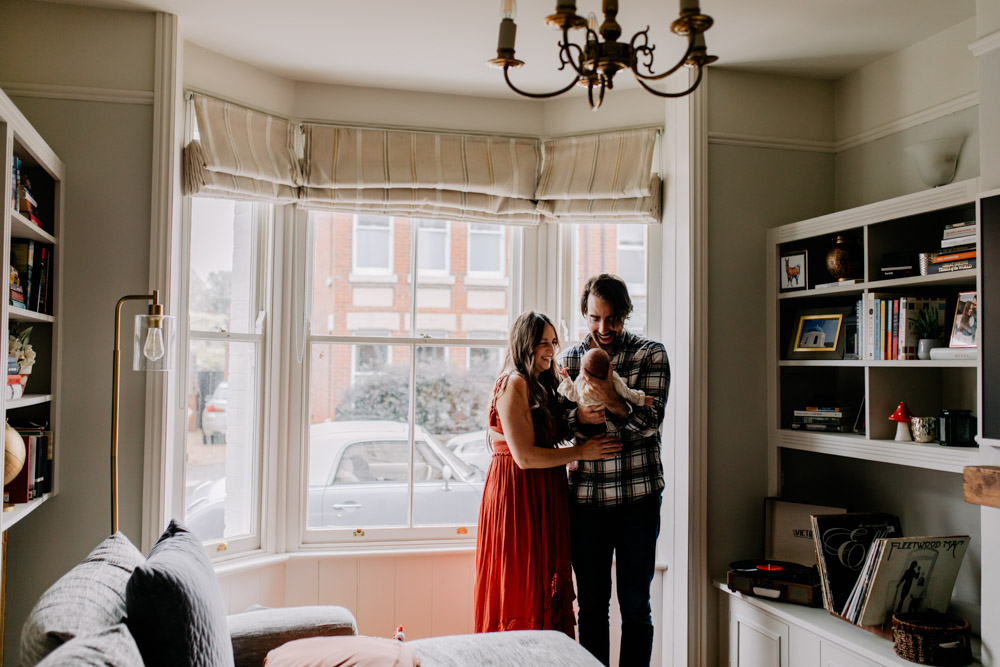 Image of new parents standing against the window and looking at their newborn baby girl. Lots of happiness and emotions. In home newborn photo sessions in Hampshire. Family photographer in Hampshire. Your Family’s Story, One Beautiful Chapter at a Time. Ewa Jones Photographer
