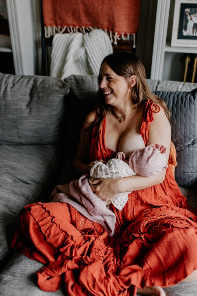 Mum is sitting on the sofa and holding newborn baby girl. Mum is wearing lovely dress. In home natural lifestyle newborn photo session in Winchester. Hampshire photographer. Ewa Jones Photography