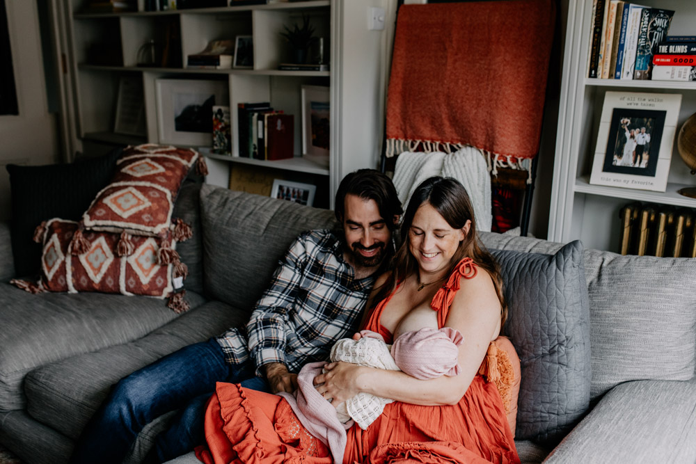 New parents are sitting on the sofa and looking down at their baby girl. Lovely in home photo shoot. What to wear for newborn photo session. Hampshire family photo shoot. Ewa Jones Photography