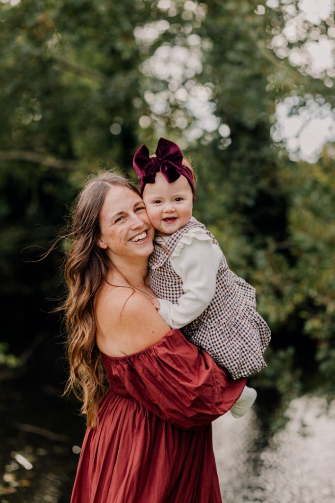 Mum is holding her one year daughter and smiling. the daughter is smiling as well and looking at the camera. both mum and daughter are wearing lovely dresses. Family photo session celebrating one year old. Outdoor family photo shoot in Hampshire. Ewa Jones Photography
