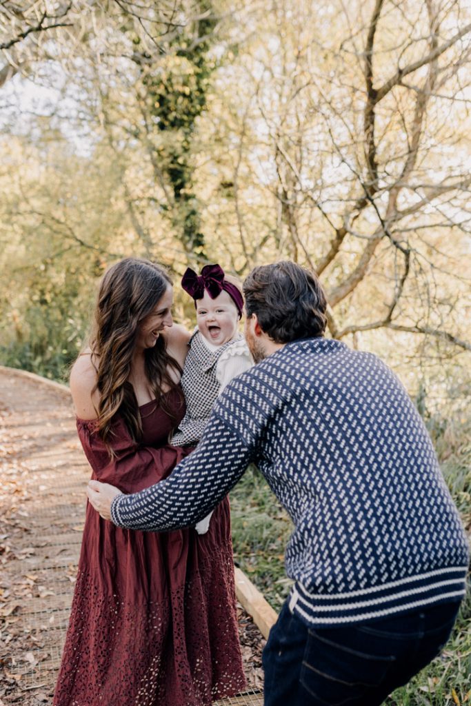 Smiling parents lift and laugh with their one-year-old baby during an outdoor family session on a wooded path, capturing a joyful, candid moment in warm natural light. Hampshire photographer. Ewa Jones Photography