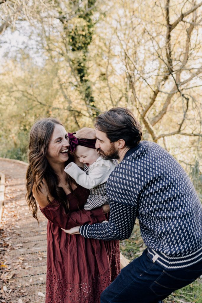 Smiling parents lift and laugh with their one-year-old baby during an outdoor family session on a wooded path, capturing a joyful, candid moment in warm natural light. Hampshire photographer. Ewa Jones Photography