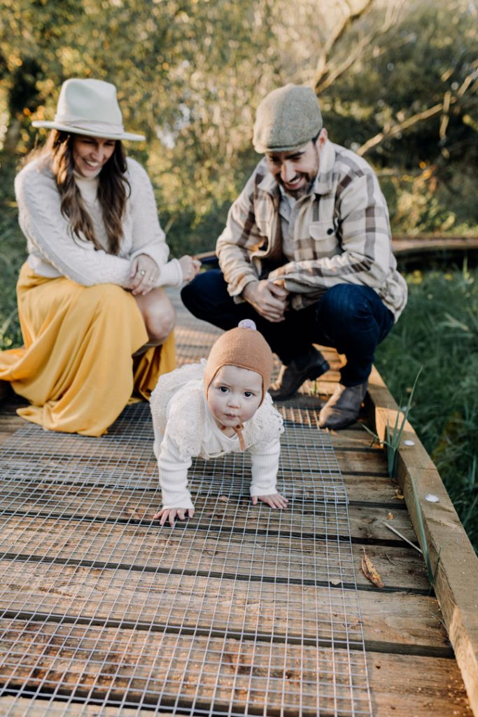 One-year-old baby crawls toward the camera on a wooden path during an outdoor family photo session, while smiling parents crouch behind, surrounded by greenery and warm natural light. Hampshire photographer. Ewa Jones Photography