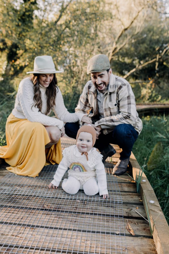One-year-old baby sits on a wooden path during an outdoor family photo session, while smiling parents crouch behind, surrounded by greenery and warm natural light. Hampshire photographer. Ewa Jones Photography