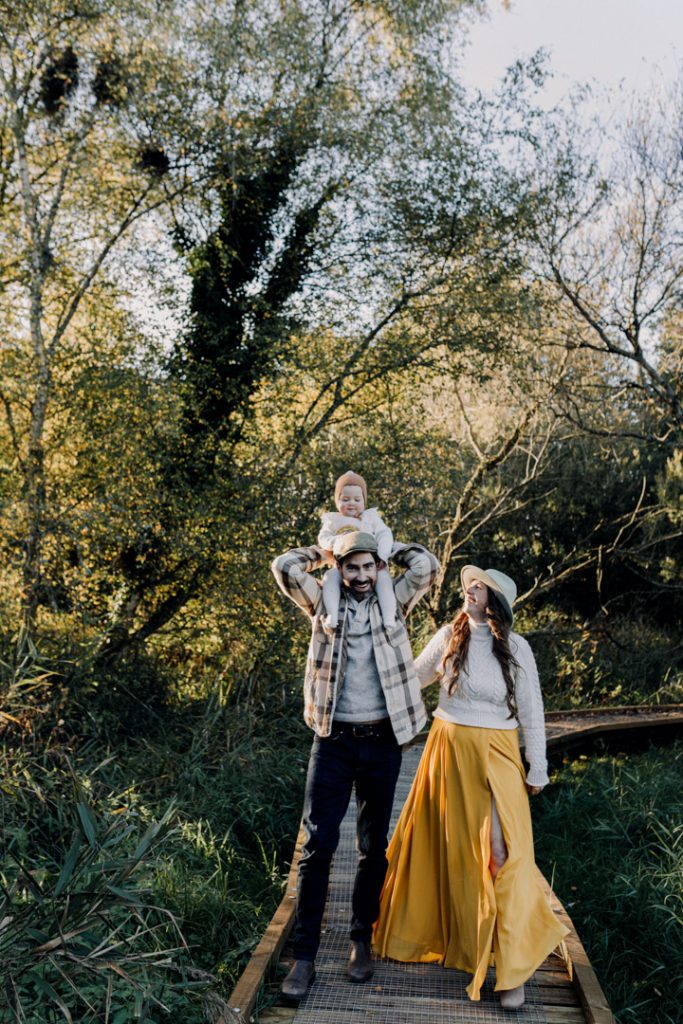One year old girl is on dad's shoulders. Mum is looking up at her daughter and smiling. Outdoor natural photoshoot during golden hour. Woodland path. outdoor session. Ewa Jones photography