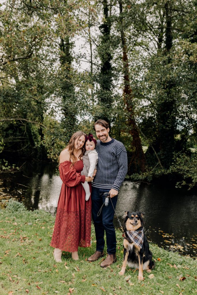 Mum and dad and their one year old girl looking at the camera and smiling. their family dog has been included in the family session. Outdoor golden hour photo session. Hampshire photo shoot. Ewa Jones Photography